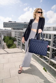 Frau auf einer Dachterrasse hält eine dunkelblaue, gesteppte Einkaufstasche in der Hand, seitliche Perspektive.