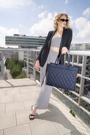 Frau auf einer Dachterrasse hält eine dunkelblaue, gesteppte Einkaufstasche in der Hand, seitliche Perspektive.