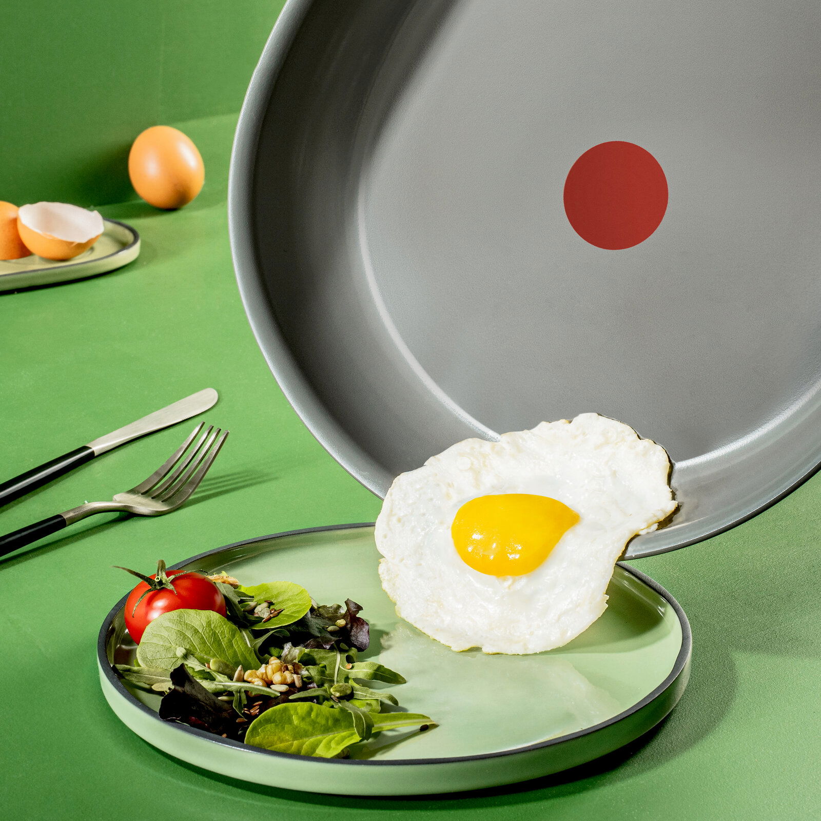 Pfanne mit grauer Antihaftbeschichtung und rotem Punkt in der Mitte, aus der ein Spiegelei auf einen Teller mit Salat gleitet. Perspektive von oben.