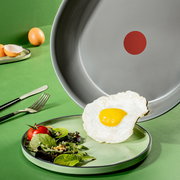 Pfanne mit grauer Antihaftbeschichtung und rotem Punkt in der Mitte, aus der ein Spiegelei auf einen Teller mit Salat gleitet. Perspektive von oben.