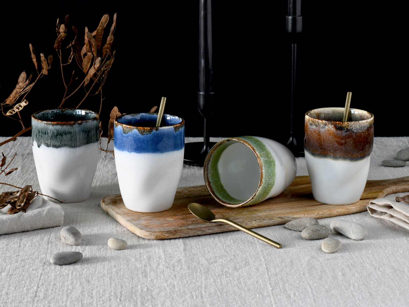 Creatable Kaffeebecher 4-tlg SQUEEZY Schräg von vorn fotografiertes Stillleben mit vier glasierten Keramikbechern in Weiß mit farbigem Rand (Grau, Blau, Grün liegend, Braun) auf Holzbrett, mit Löffel, Steinen und Trockenblumen auf Leinentuch vor dunklem Hintergrund