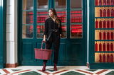 Frau in schwarzem Outfit steht vor einer grünen Tür und hält eine rote Tragetasche in der Hand, Frontansicht.