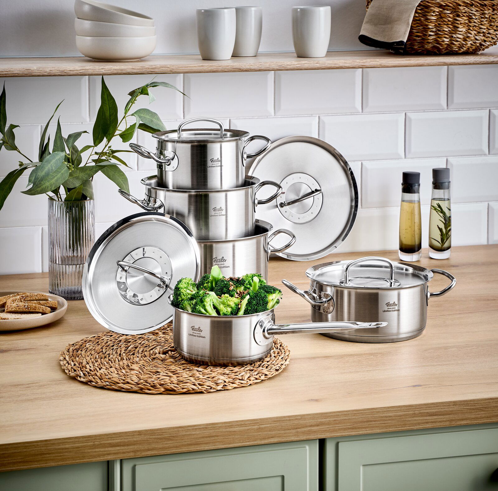 Stapel von Edelstahlkochtöpfen auf einer Küchenarbeitsplatte, mit einem Topf voller Brokkoli im Vordergrund, seitliche Perspektive.