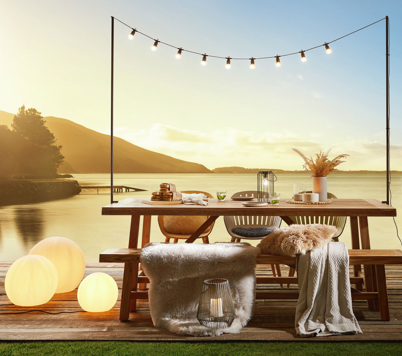 Gartentisch und Gartenbank auf einer Terrasse mit Seeblick bei Sonnenuntergang, dekoriert mit Lichtern und Kissen, seitliche Perspektive