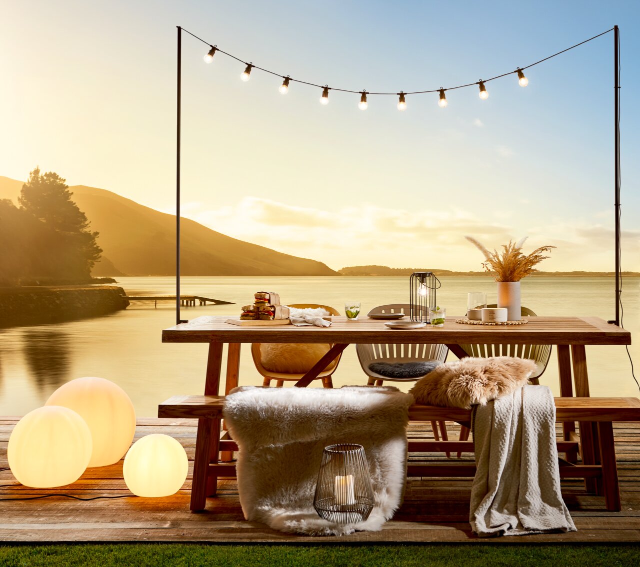Gartentisch und Gartenbank auf einer Terrasse mit Seeblick bei Sonnenuntergang, dekoriert mit Lichtern und Kissen, seitliche Perspektive