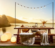 Gartentisch und Gartenbank auf einer Terrasse mit Seeblick bei Sonnenuntergang, dekoriert mit Lichtern und Kissen, seitliche Perspektive