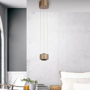 Moderne zylindrische Pendelleuchte in Messingoptik mit langem Kabel, hängend vor heller Betonwand im Wohnraum, frontal aus leicht erhöhter Perspektive