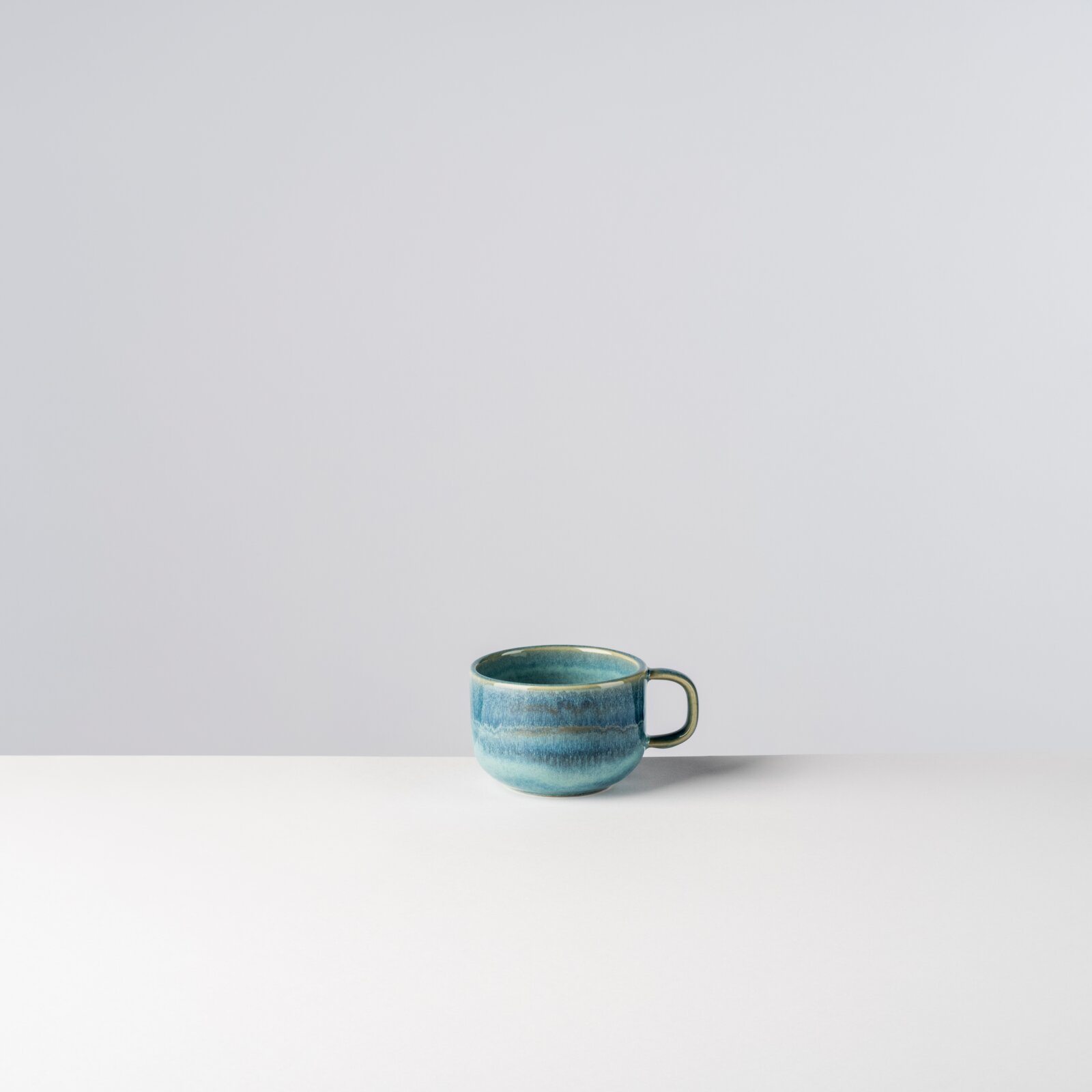 Blau glasierte Keramik-Kaffeetasse mit Henkel auf weißer Fläche vor hellem Hintergrund, Frontansicht leicht von oben