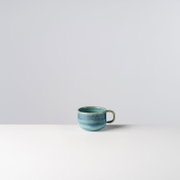 Blau glasierte Keramik-Kaffeetasse mit Henkel auf weißer Fläche vor hellem Hintergrund, Frontansicht leicht von oben