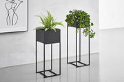 Zwei moderne, schwarze Blumensäulen mit Pflanzen, auf einem hellen Boden vor einer weißen Wand. Perspektive von vorne.
