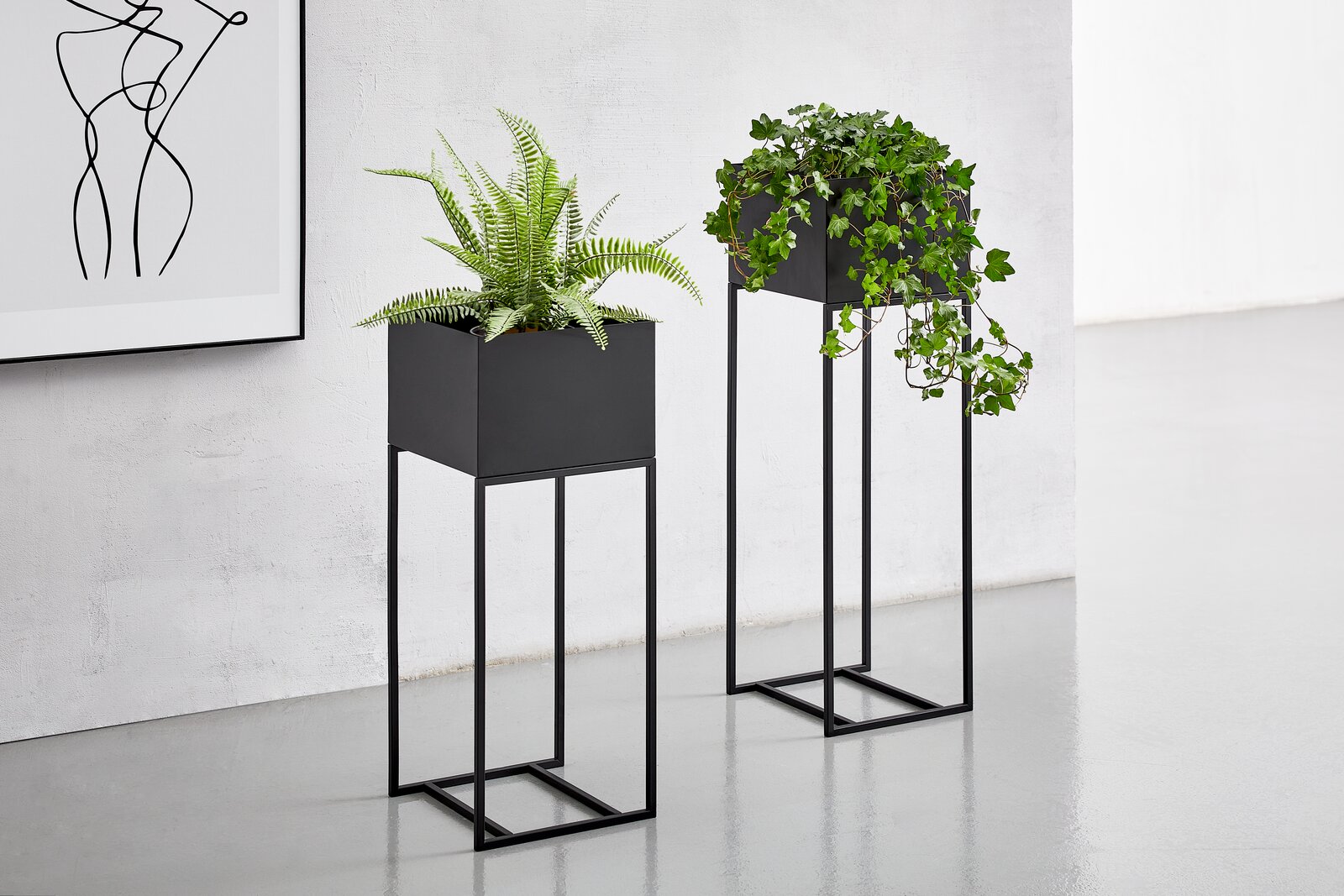 Zwei moderne, schwarze Blumensäulen mit Pflanzen, auf einem hellen Boden vor einer weißen Wand. Perspektive von vorne.