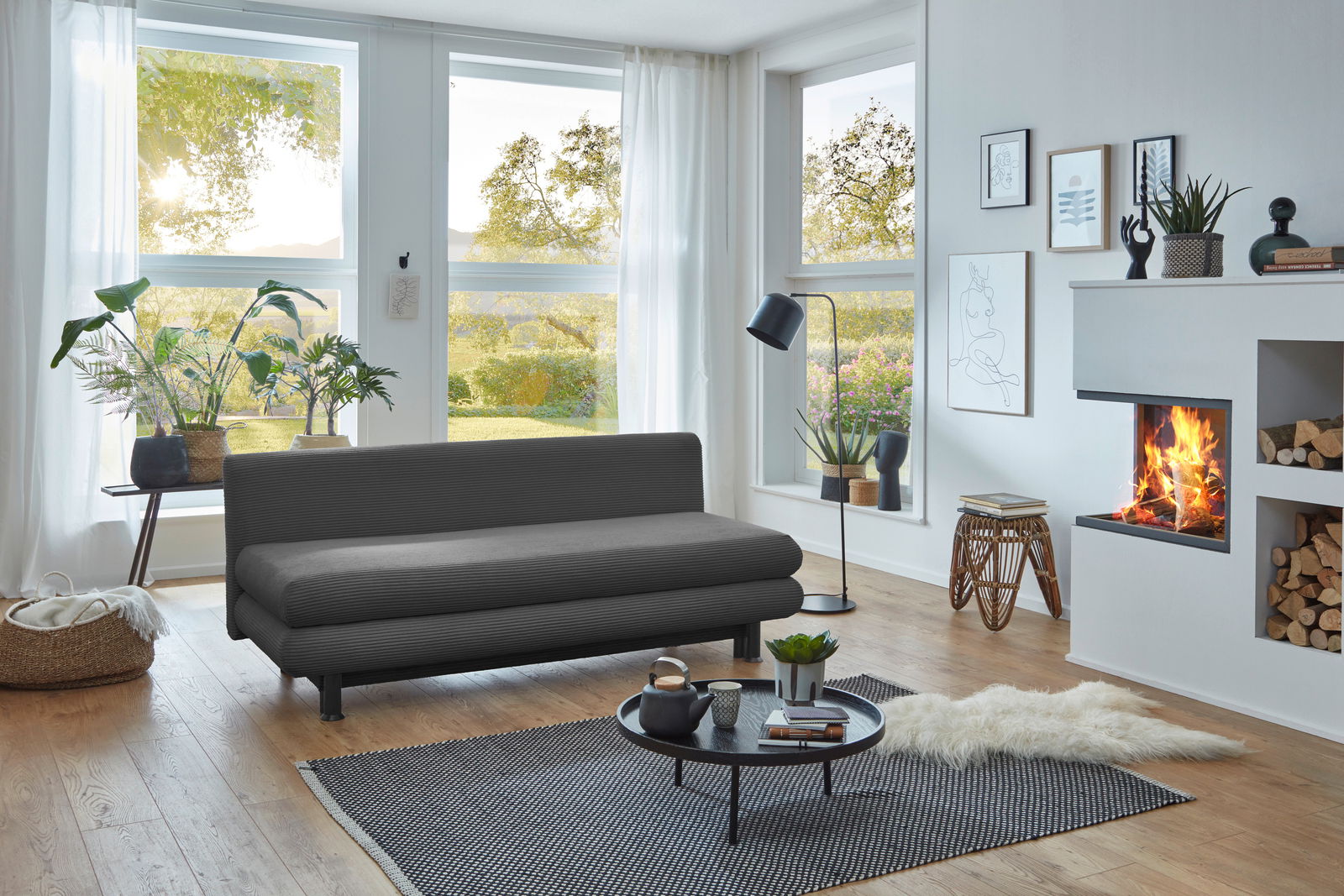 Graues Funktionssofa in modernem Wohnzimmer mit großen Fenstern, Pflanzen und Kamin, seitliche Perspektive.