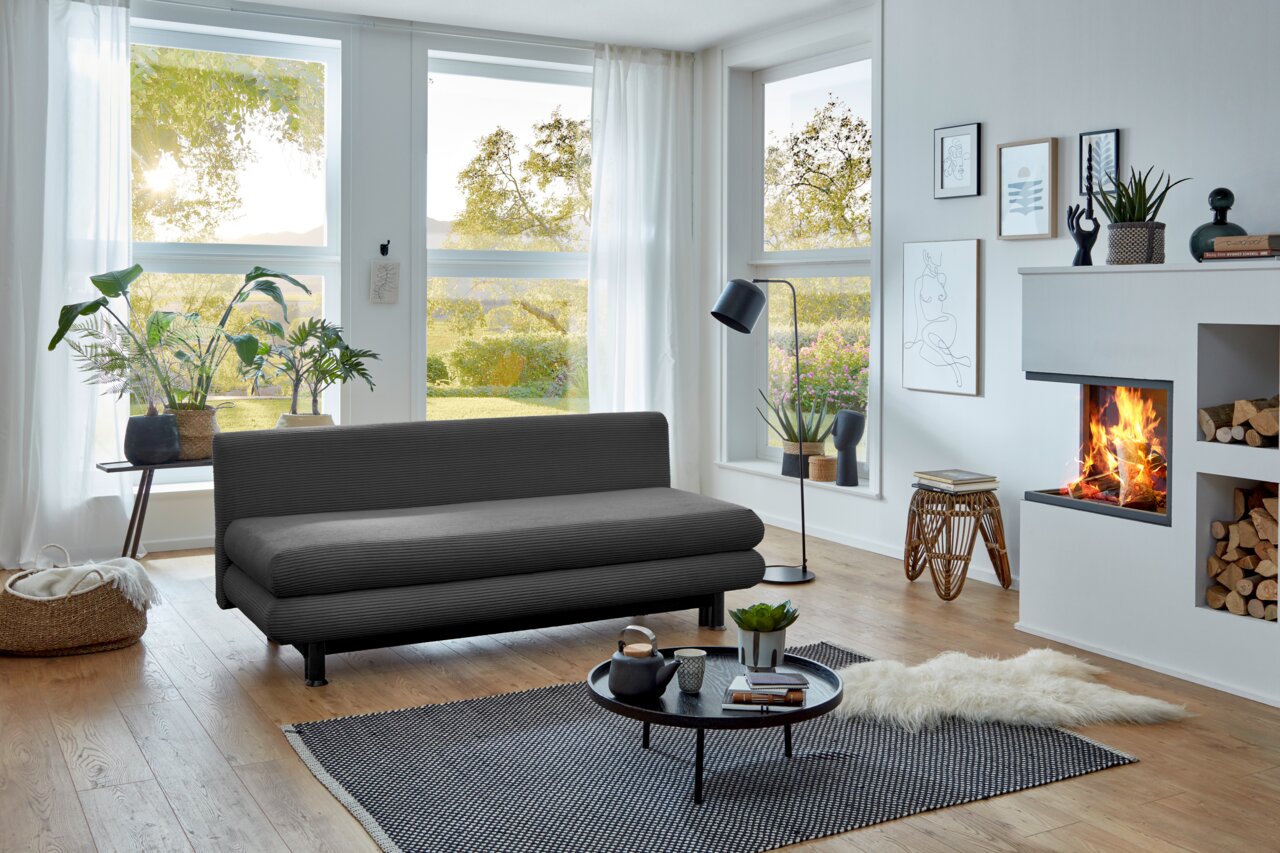 Graues Funktionssofa in modernem Wohnzimmer mit großen Fenstern, Pflanzen und Kamin, seitliche Perspektive.