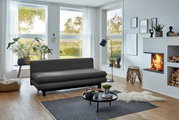Graues Funktionssofa in modernem Wohnzimmer mit großen Fenstern, Pflanzen und Kamin, seitliche Perspektive.