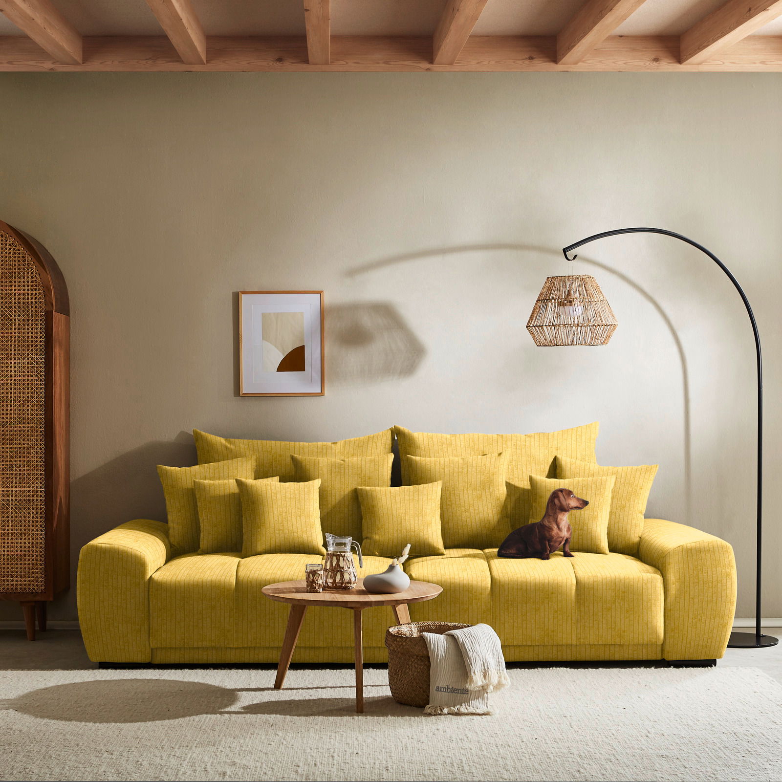 Großes gelbes Sofa mit vielen Kissen, ein Hund sitzt darauf, im Hintergrund eine Stehlampe und ein Bild an der Wand, Perspektive von vorne.