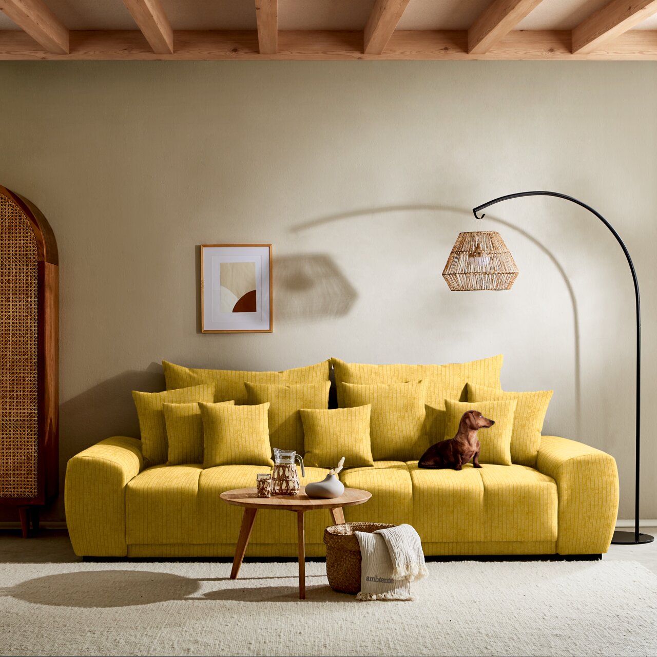 Großes gelbes Sofa mit vielen Kissen, ein Hund sitzt darauf, im Hintergrund eine Stehlampe und ein Bild an der Wand, Perspektive von vorne.