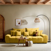 Großes gelbes Sofa mit vielen Kissen, ein Hund sitzt darauf, im Hintergrund eine Stehlampe und ein Bild an der Wand, Perspektive von vorne.