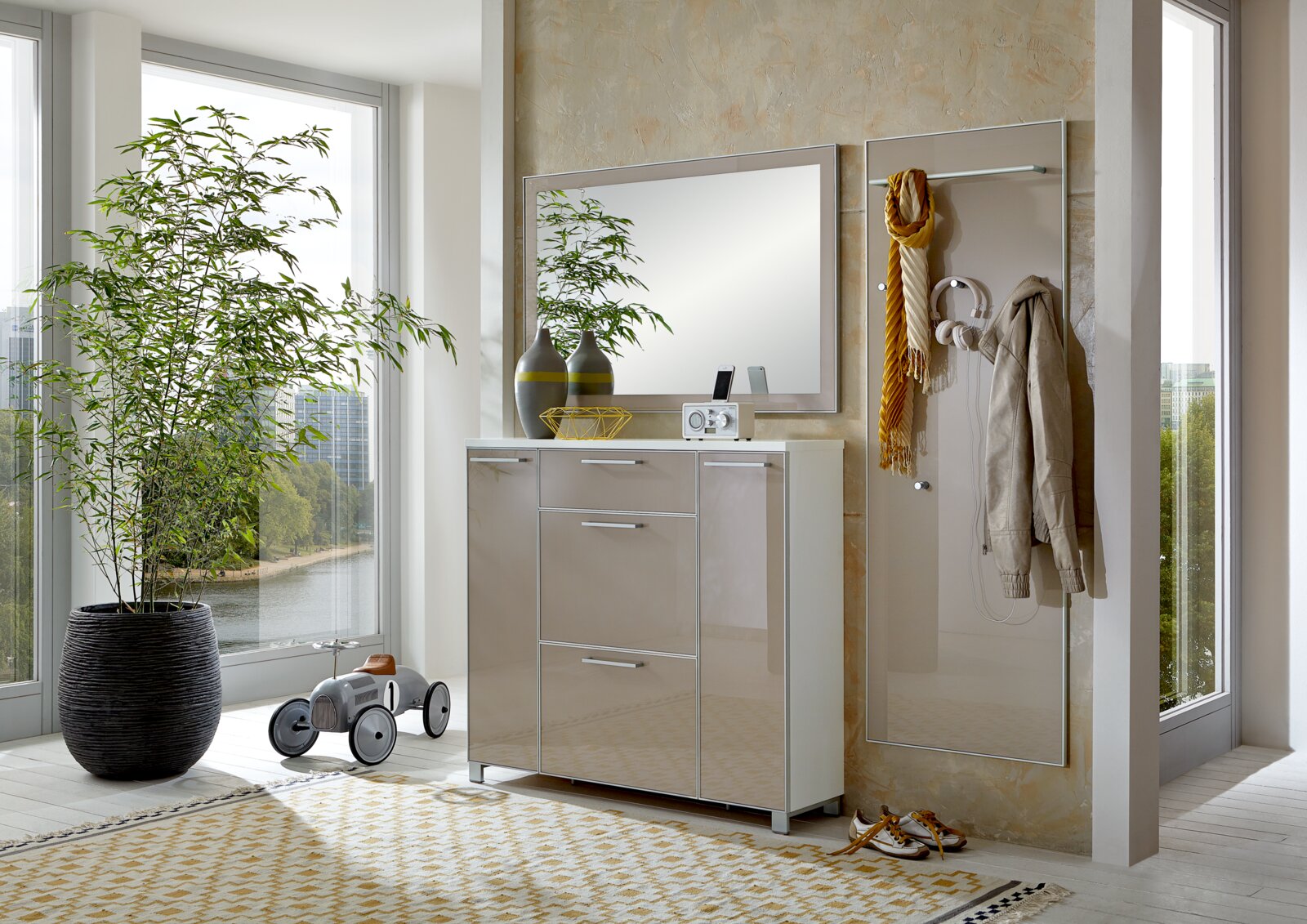 Moderne garderobeset in een lichte ruimte met een groot raam, bestaande uit een kast met hoogglansfronten, een spiegel en een wandgarderobe met haken. Aanzicht van voren.
