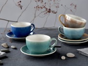 Stillleben mit glasierten Keramik-Kaffeetassen mit Untertassen in Blau und Türkis sowie gestapelten Tassen mit goldfarbenem Henkel auf grauem Tisch, perspektivisch aus leicht erhöhter Frontansicht.