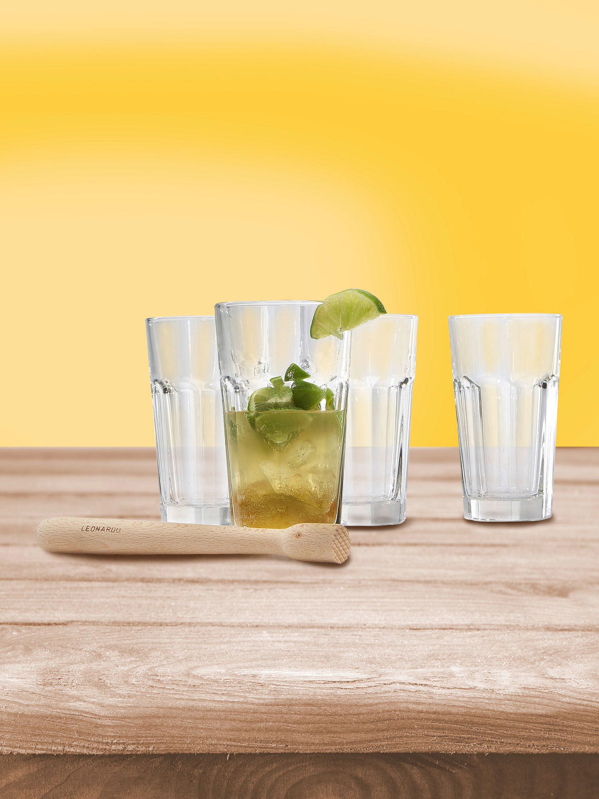 Vier Gläser und ein Holzstößel auf einem Holztisch vor gelbem Hintergrund, frontale Perspektive