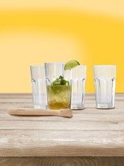 Vier Gläser und ein Holzstößel auf einem Holztisch vor gelbem Hintergrund, frontale Perspektive