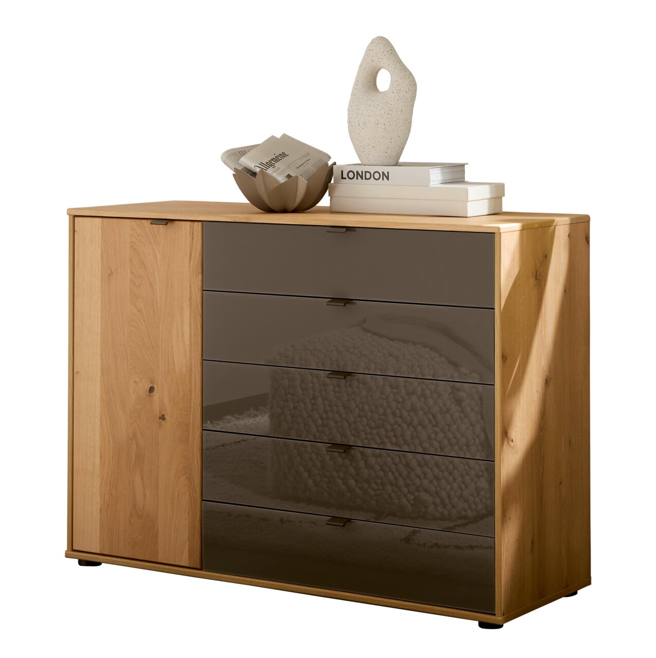 Linea Natura Kommode CARDIFF Moderne Kommode aus hellem Holz mit linker Tür und fünf rauchgrauen Glas-Schubladen, dekoriert mit Büchern und Vase, in frontaler leicht rechter Perspektive.