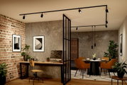 Modernes Industrial-Interieur mit schwarzer Schienenbeleuchtung und Spots an der Decke, Blick aus leichtem Untersicht-/Weitwinkelwinkel in Arbeits- und Essbereich mit Ziegel- und Betonwand, Glasraumteiler, Holzschreibtisch und Esstisch mit braunen Stühlen