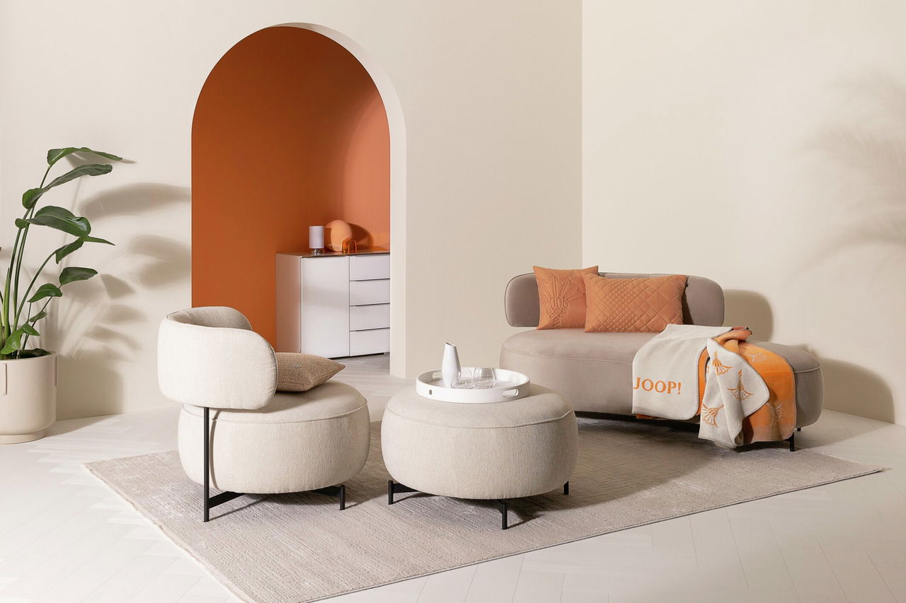 JOOP! Zierkissenhülle J!MOVE Modernes Wohnzimmer mit beigem Sofa und zwei passenden Sesseln, dekoriert mit orangefarbenen Zierkissen und einer Decke, aus der Perspektive von vorne.
