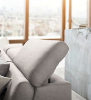 Nahaufnahme einer modernen, hellgrauen Rundecke mit verstellbarer Kopfstütze in einem hellen Raum mit großen Fenstern im Hintergrund.