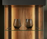 Nahaufnahme einer beleuchteten Vitrine mit zwei modernen, geometrischen Gläsern auf einem Glasregal, Holzrückwand im Hintergrund.