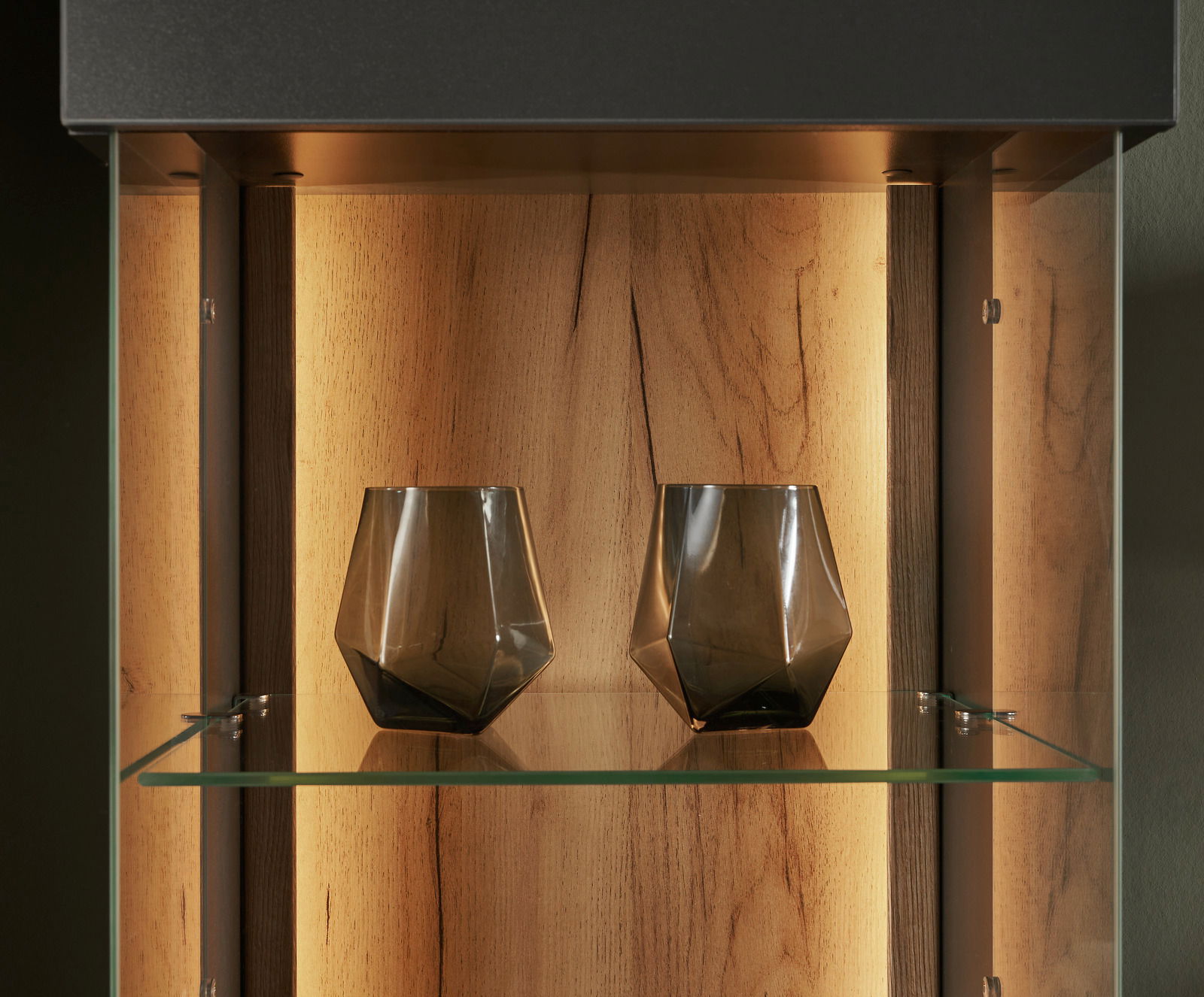 Nahaufnahme einer beleuchteten Vitrine mit zwei modernen, geometrischen Gläsern auf einem Glasregal, Holzrückwand im Hintergrund.