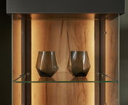 Nahaufnahme einer beleuchteten Vitrine mit zwei modernen, geometrischen Gläsern auf einem Glasregal, Holzrückwand im Hintergrund.