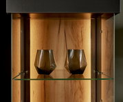 Close-up van een verlichte vitrine met twee moderne, geometrische glazen op een glazen plank, met een houten achterwand op de achtergrond.