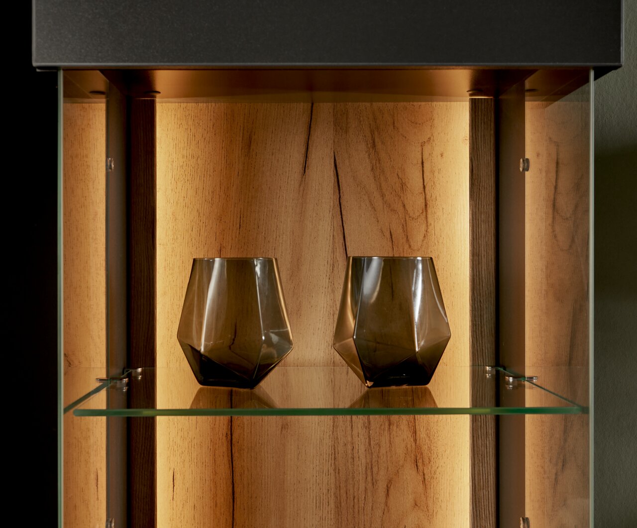 Close-up van een verlichte vitrine met twee moderne, geometrische glazen op een glazen plank, met een houten achterwand op de achtergrond.