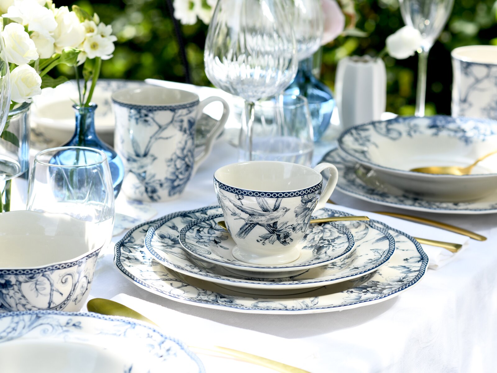 Schräg von vorne fotografiertes, elegant gedecktes Tischarrangement mit blau-weiß gemustertem Porzellan (Tasse auf Untertasse und Teller), goldfarbenem Besteck, Weingläsern und Blumen in blauer Vase auf weißer Tischdecke.