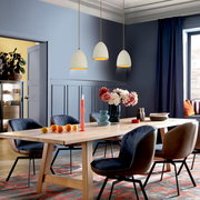 Modernes Esszimmer mit langem Holzesstisch und gepolsterten Stühlen, dekoriert mit Blumen, Kerzen und Geschirr; darüber drei cremefarbene Pendelleuchten, fotografiert aus schräger Frontperspektive auf Tischhöhe.
