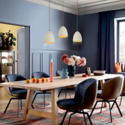 Modernes Esszimmer mit langem Holzesstisch und gepolsterten Stühlen, dekoriert mit Blumen, Kerzen und Geschirr; darüber drei cremefarbene Pendelleuchten, fotografiert aus schräger Frontperspektive auf Tischhöhe.