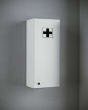 Weißer Erste-Hilfe-Wandschrank mit schwarzem Kreuzsymbol und Schloss, an einer grauen Wand montiert, schräg von vorn links aus leicht erhöhter Perspektive