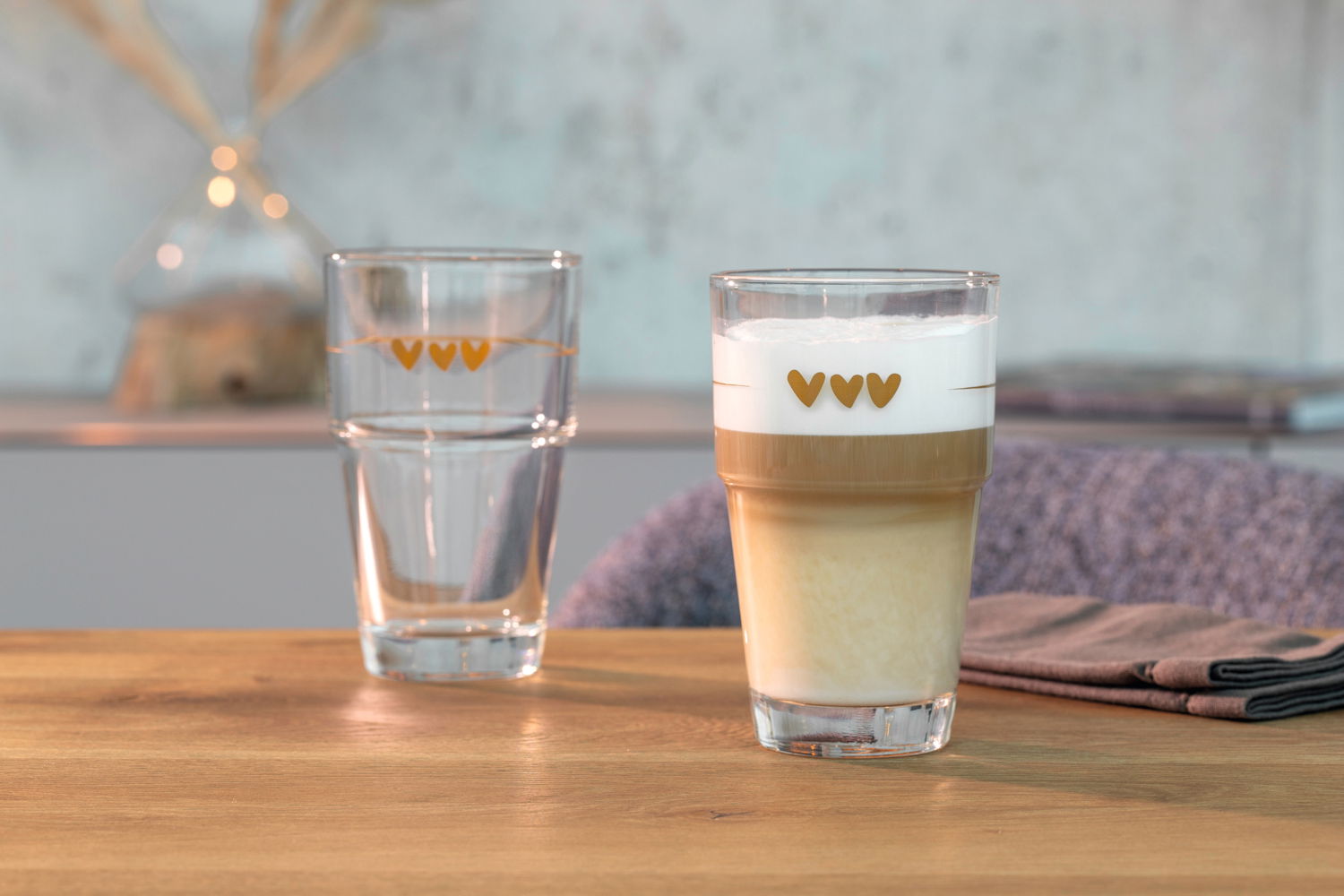 Zwei Gläser mit Herzmotiven, eines mit Latte Macchiato gefüllt, auf einem Holztisch. Vorderansicht.