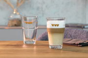 Zwei Gläser mit Herzmotiven, eines mit Latte Macchiato gefüllt, auf einem Holztisch. Vorderansicht.