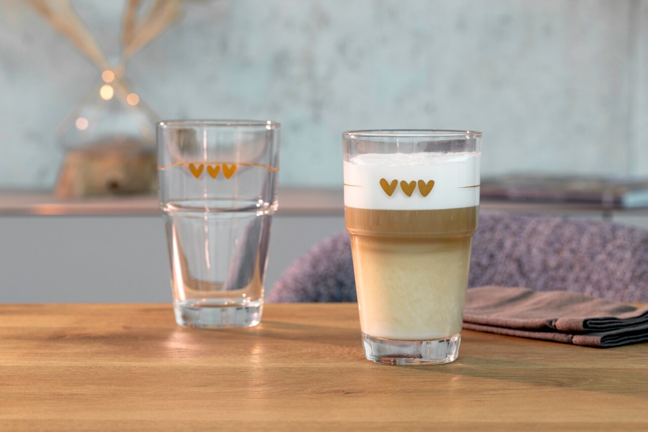 Twee glazen met hartjesmotieven, waarvan één gevuld met latte macchiato, op een houten tafel. Vooraanzicht.