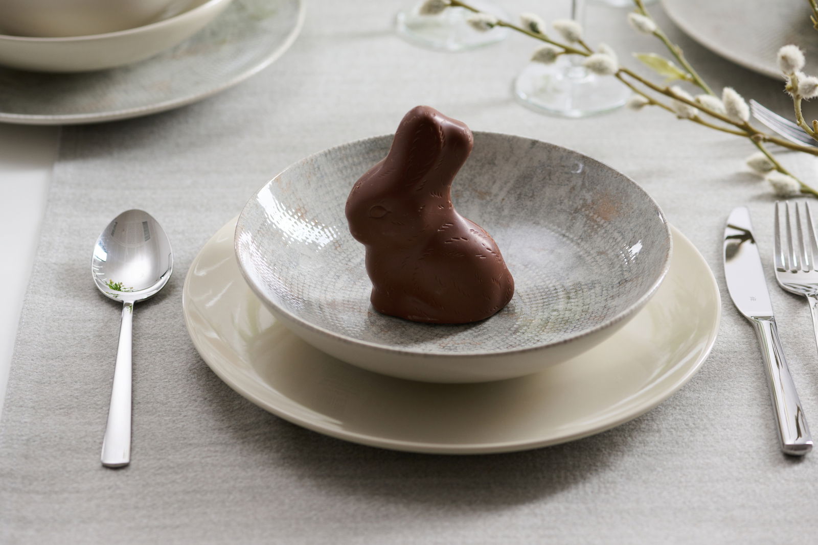 Ein gedeckter Tisch mit einem Schokoladenhasen in einer Schale auf einem Teller, umgeben von Besteck und einem dezenten Tischläufer. Perspektive von oben.