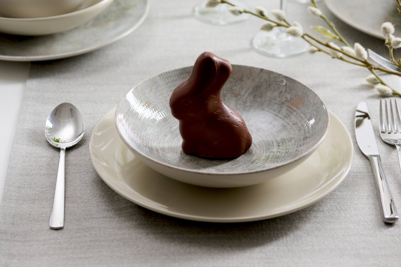 Ein gedeckter Tisch mit einem Schokoladenhasen in einer Schale auf einem Teller, umgeben von Besteck und einem dezenten Tischläufer. Perspektive von oben.
