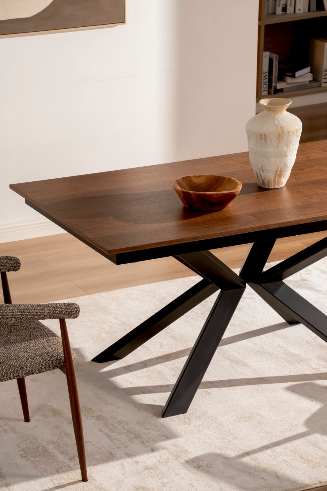 Moderne eettafel met donkerbruin houten tafelblad en zwart metalen kruispootonderstel, gestyled met een houten schaal en een witte vaas; perspectief schuin van voren op tafelhoogte.