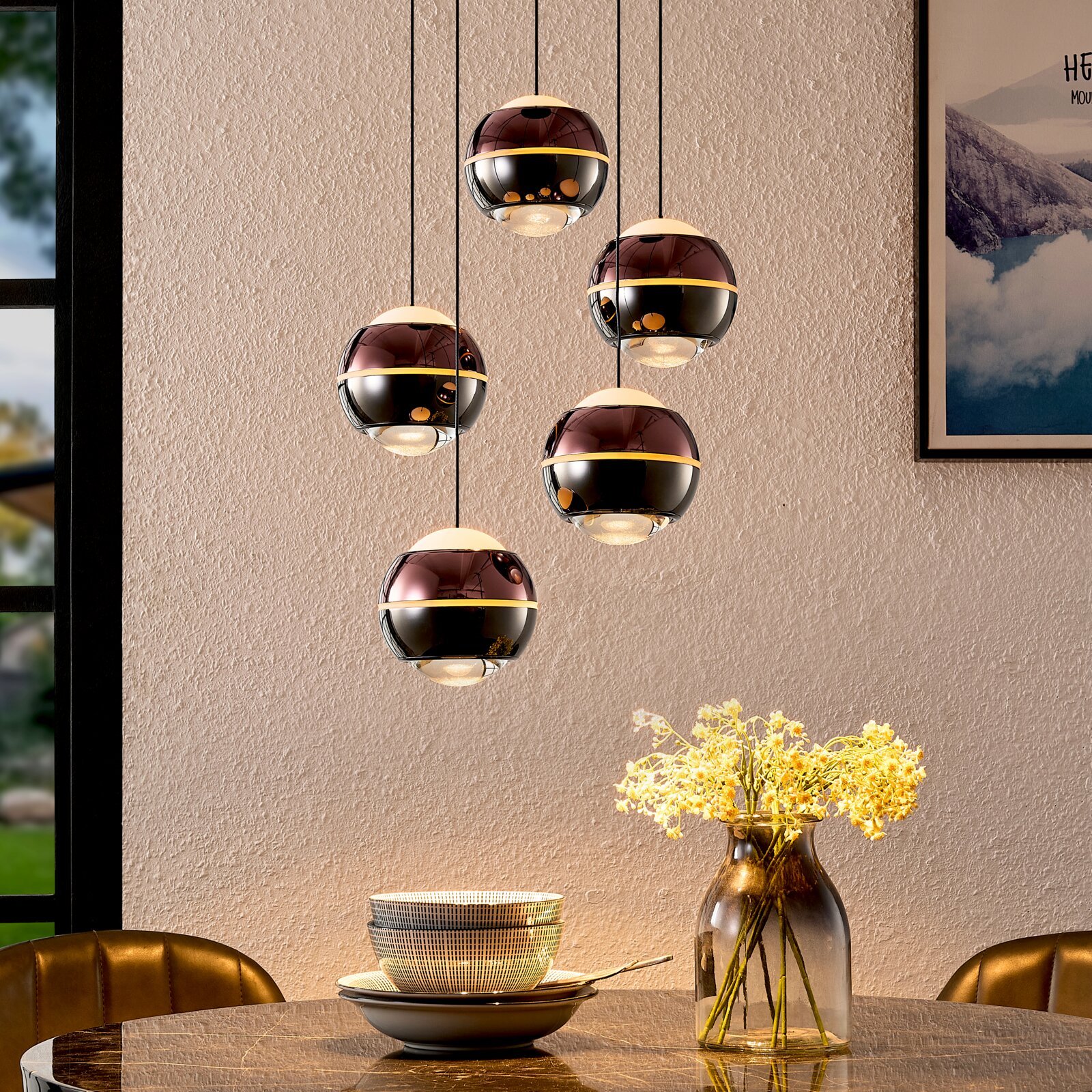 Moderne Pendelleuchte mit fünf kugelförmigen, rauchglasfarbenen Lampenschirmen und goldfarbenem Ring, hängend über einem Esstisch mit Vase; Perspektive: frontale Innenraumansicht leicht von unten.