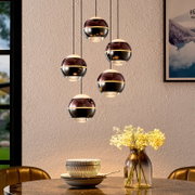 Moderne Pendelleuchte mit fünf kugelförmigen, rauchglasfarbenen Lampenschirmen und goldfarbenem Ring, hängend über einem Esstisch mit Vase; Perspektive: frontale Innenraumansicht leicht von unten.