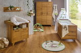 Komplettes Babyzimmer-Set aus Holz mit Babybett, Wickelkommode, 3-türigem Kleiderschrank und Wandregal, aus der Perspektive eines hellen Raumes mit großen Fenstern.