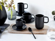 Mattschwarzes Tassen- und Untertassen-Set in verschiedenen Größen, aus leicht erhöhter Frontalperspektive stilvoll auf einem hellen Tisch mit Holzbrett arrangiert
