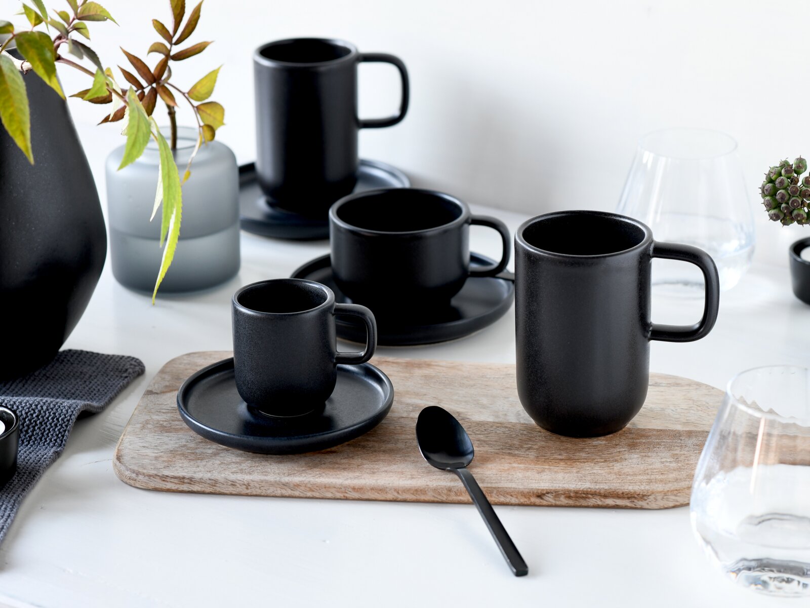 Mattschwarzes Tassen- und Untertassen-Set in verschiedenen Größen, aus leicht erhöhter Frontalperspektive stilvoll auf einem hellen Tisch mit Holzbrett arrangiert