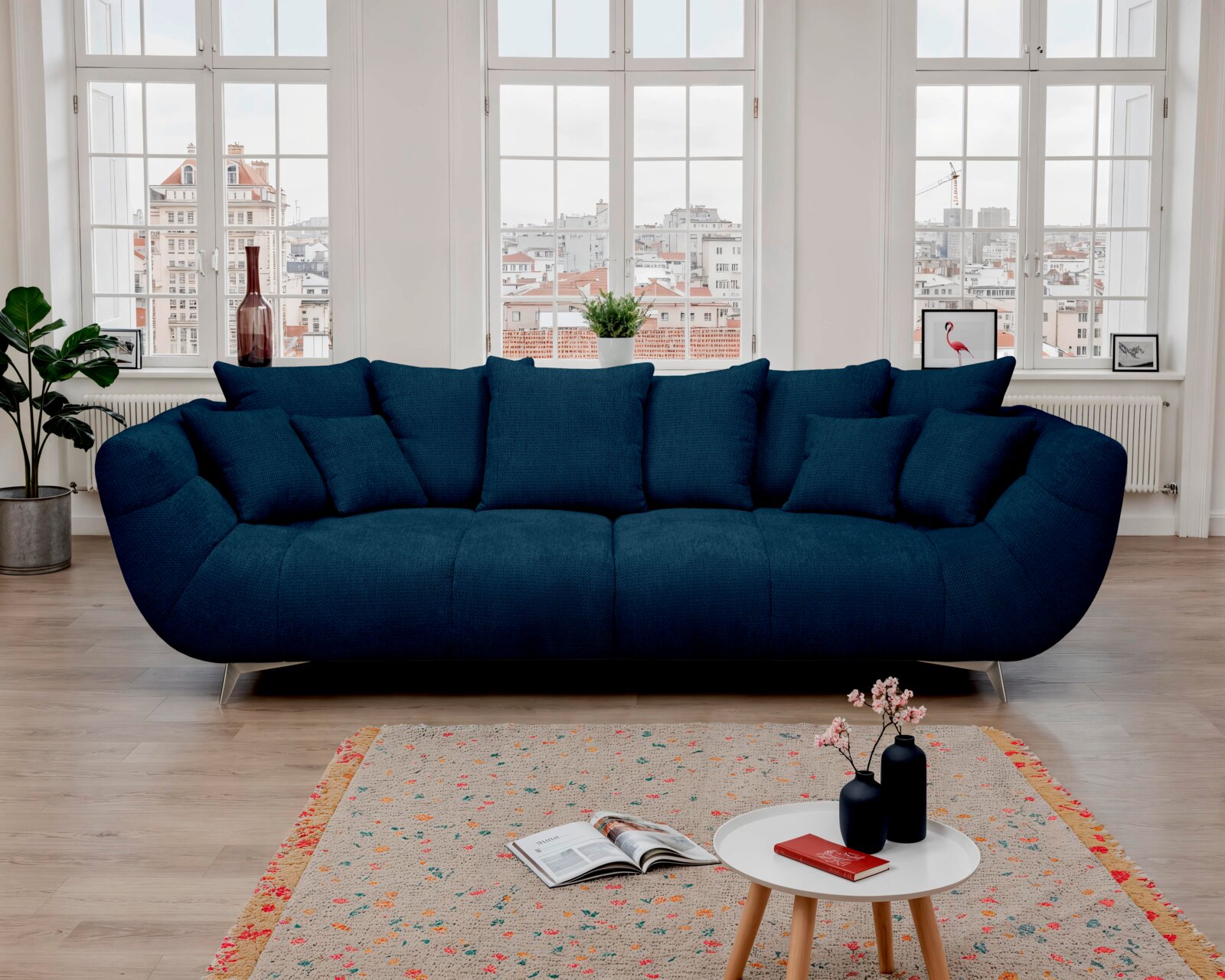 Dunkelblaues, großzügig gepolstertes 3-Sitzer-Sofa mit mehreren Kissen und schrägen Metallfüßen im hellen Wohnzimmer vor großen Fenstern, frontal aus Augenhöhe fotografiert.